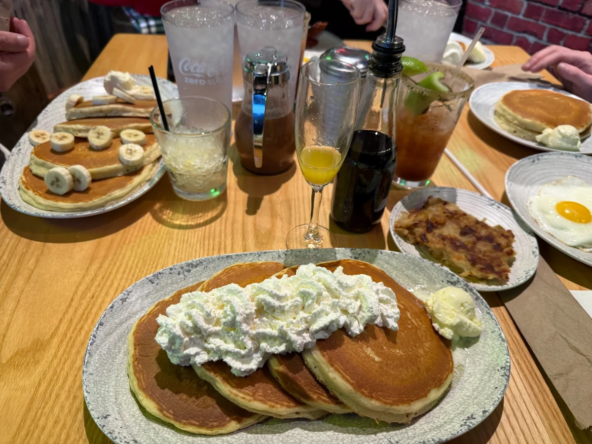 A breakfast table with pancakes topped with whipped cream, pancakes with sliced bananas, a sunny-side-up egg, hash browns, drinks, and condiments on a wooden surface.