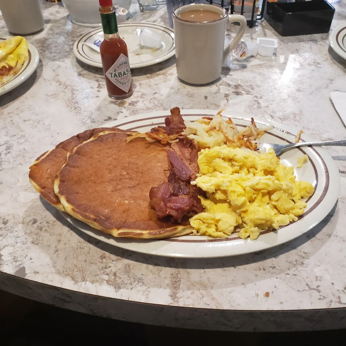 A breakfast plate with pancakes, scrambled eggs, bacon, and hash browns sits on a marble table next to a mug of coffee and a bottle of Tabasco sauce.