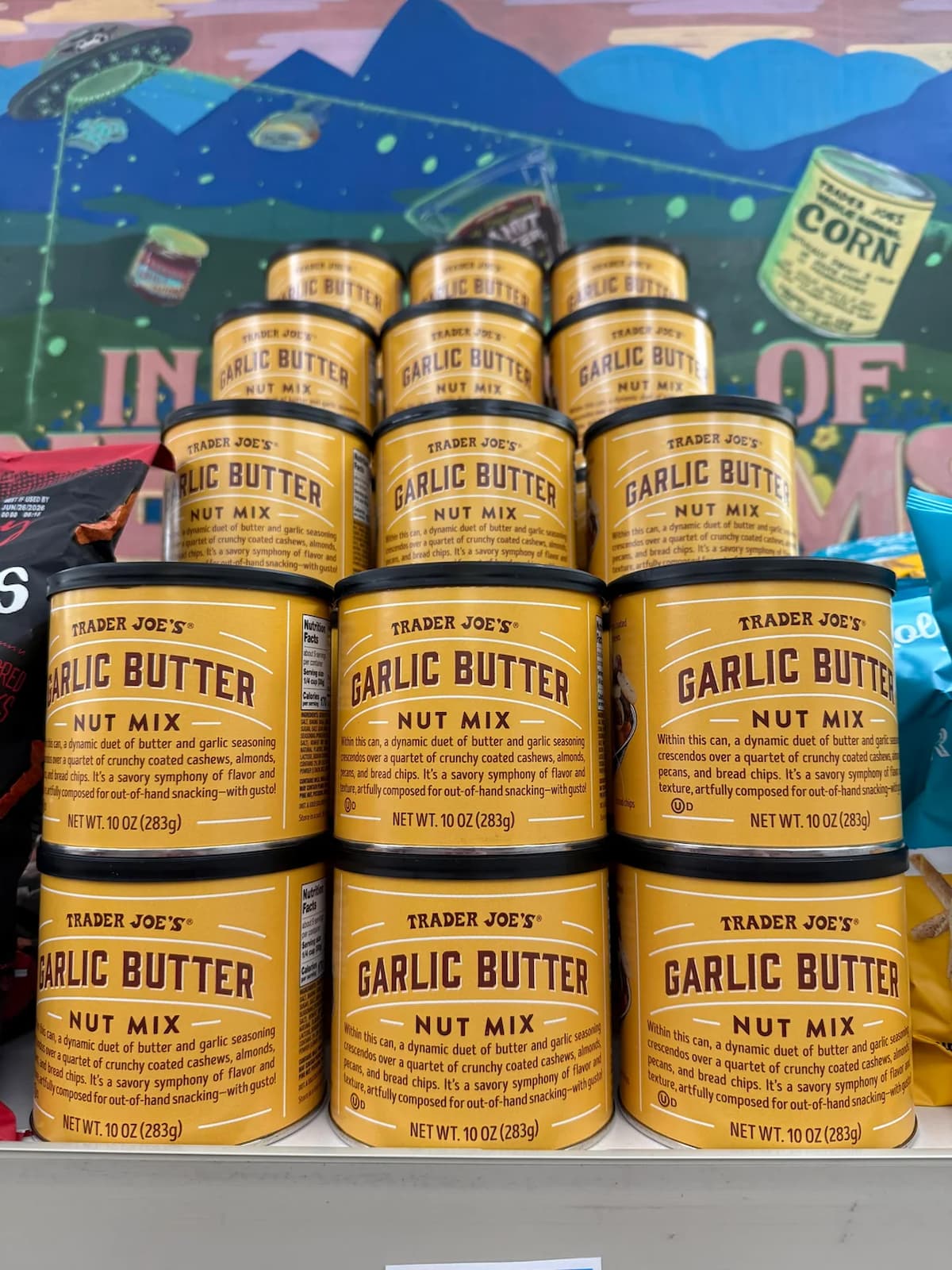 A display of Trader Joe's Garlic Butter Nut Mix cans are stacked in a pyramid shape on a store shelf, with colorful packaging and a mural visible in the background.