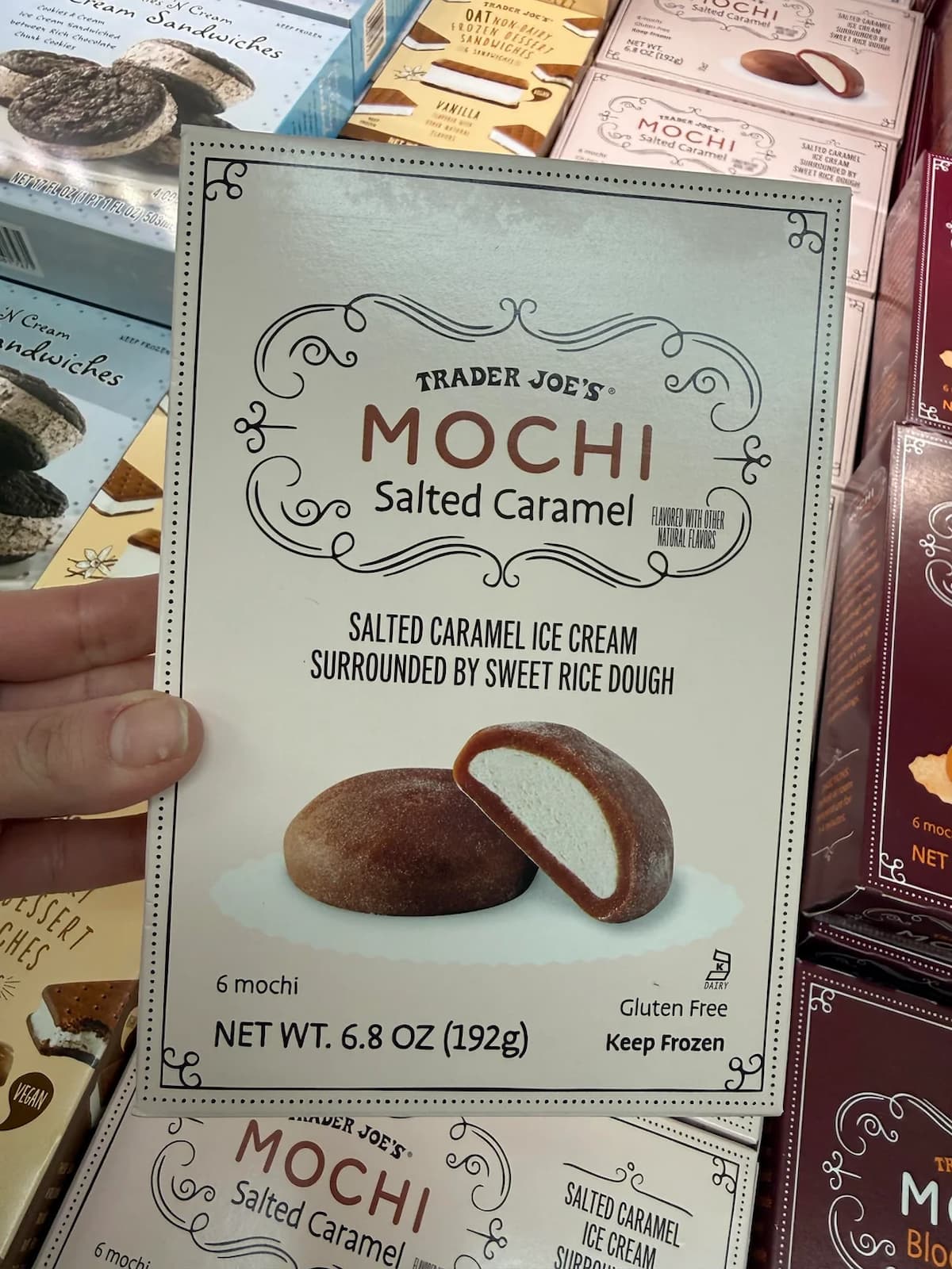 A hand holds a box of Trader Joe's Salted Caramel Mochi, featuring an image of two mochi pieces with caramel ice cream inside sweet rice dough. The box states it contains 6 mochi, is gluten-free, and should be kept frozen.