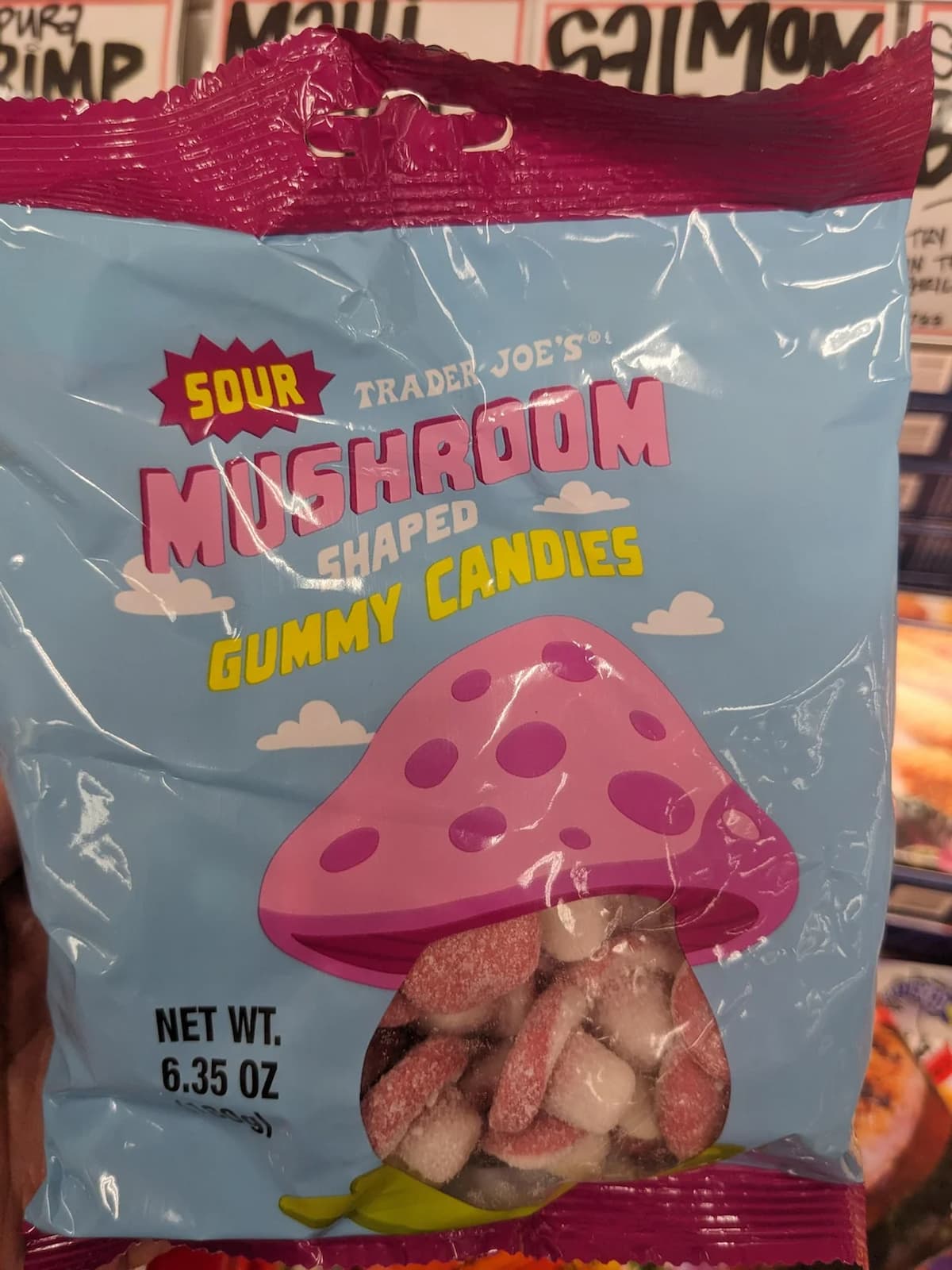 A blue bag of Trader Joe's Sour Mushroom Shaped Gummy Candies with a large pink-spotted cartoon mushroom illustration on the front. The bag is partly transparent, showing red and white gummies inside. Net weight: 6.35 oz.