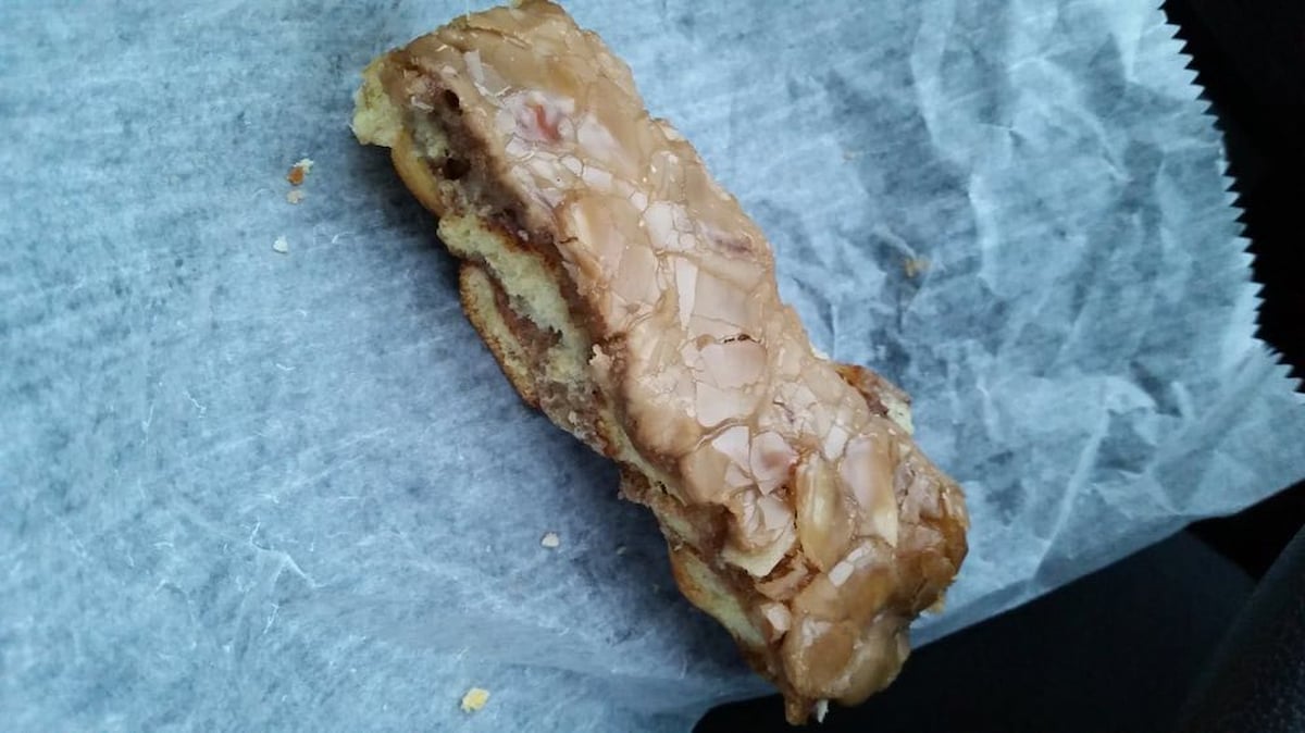 A rectangular pastry with a shiny, glazed topping sits on a sheet of crinkled wax paper. The pastry is slightly broken at one end, revealing a light and airy interior.