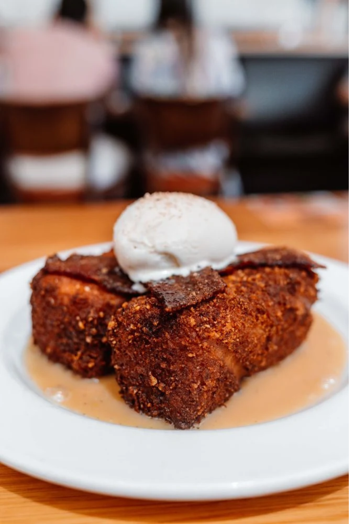 A thick slice of golden-brown bread pudding topped with a scoop of vanilla ice cream and caramel sauce, served on a white plate with a blurred background of people sitting at a table.
