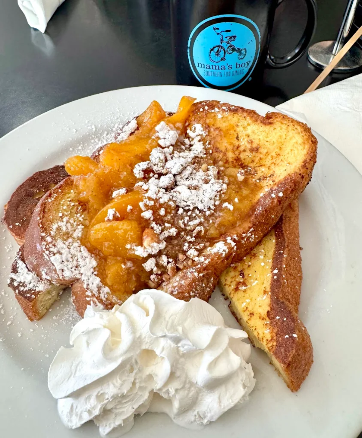 French toast topped with peach compote and powdered sugar, served with a generous dollop of whipped cream on a white plate. A black mug labeled "mama's boy" is in the background.