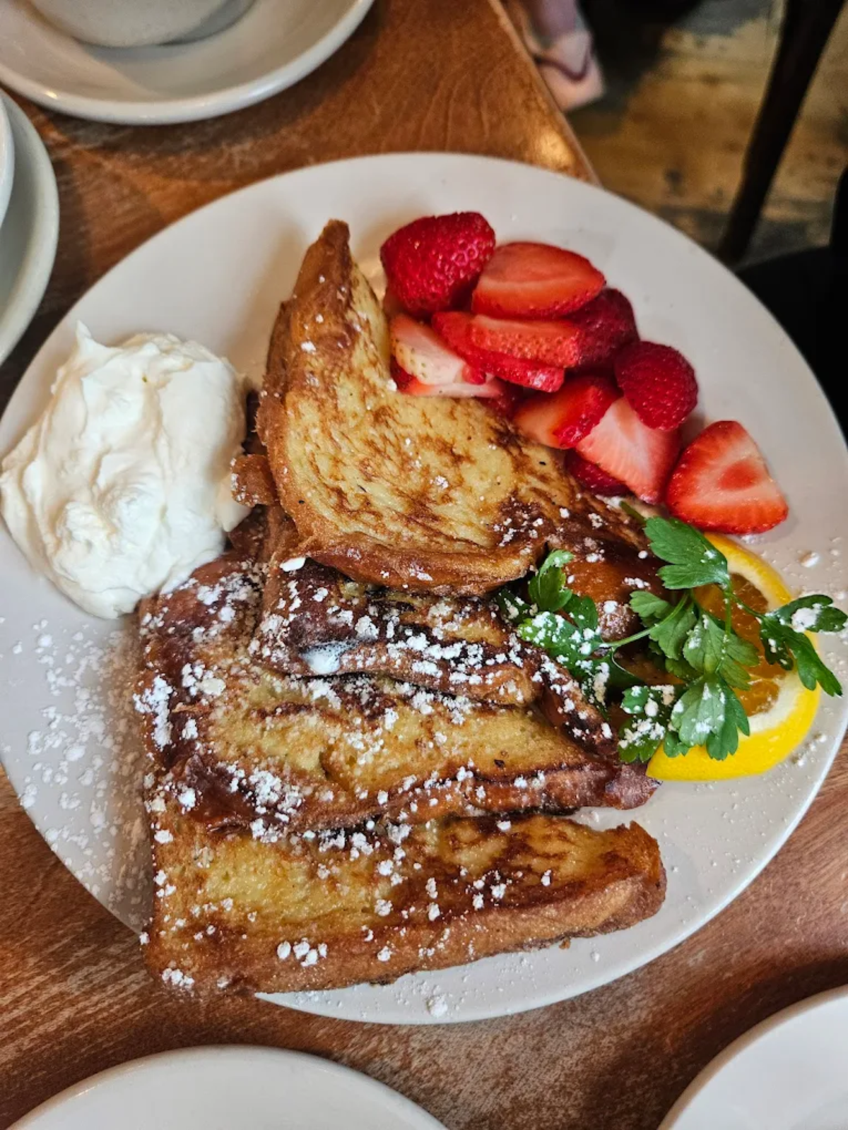 A plate with three slices of French toast dusted with powdered sugar, whipped cream on the side, sliced strawberries, and a garnish of orange and parsley.