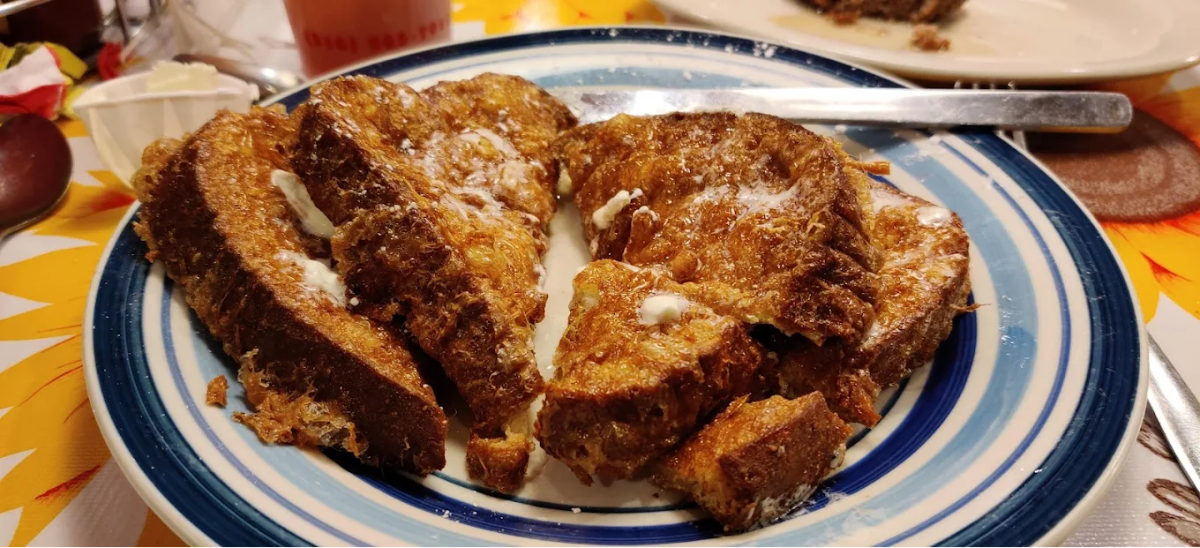 A plate of French toast slices topped with butter is served on a blue and white striped dish, with a knife and a small dish of cream or butter nearby on a sunflower-patterned tablecloth.