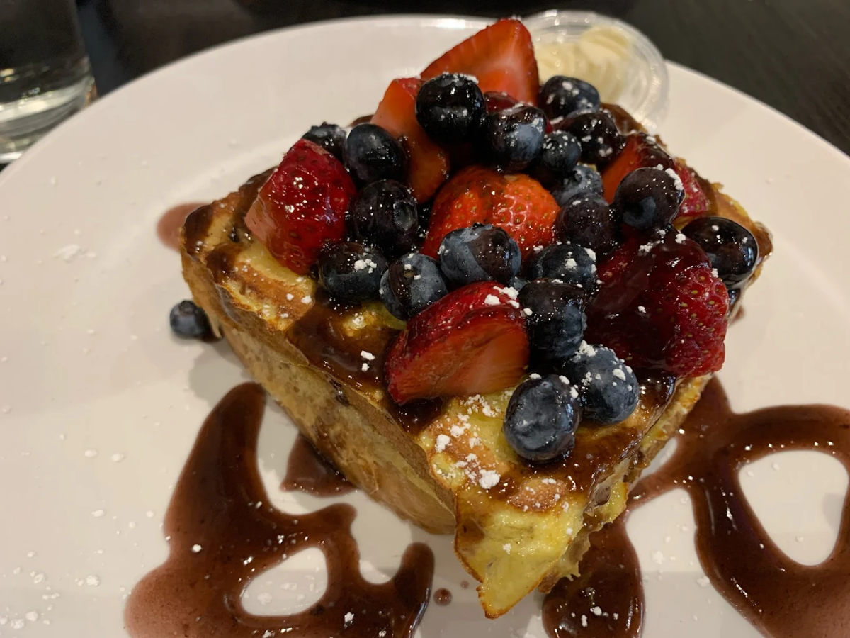 A slice of thick French toast topped with fresh strawberries and blueberries, drizzled with chocolate sauce, and sprinkled with powdered sugar on a white plate.