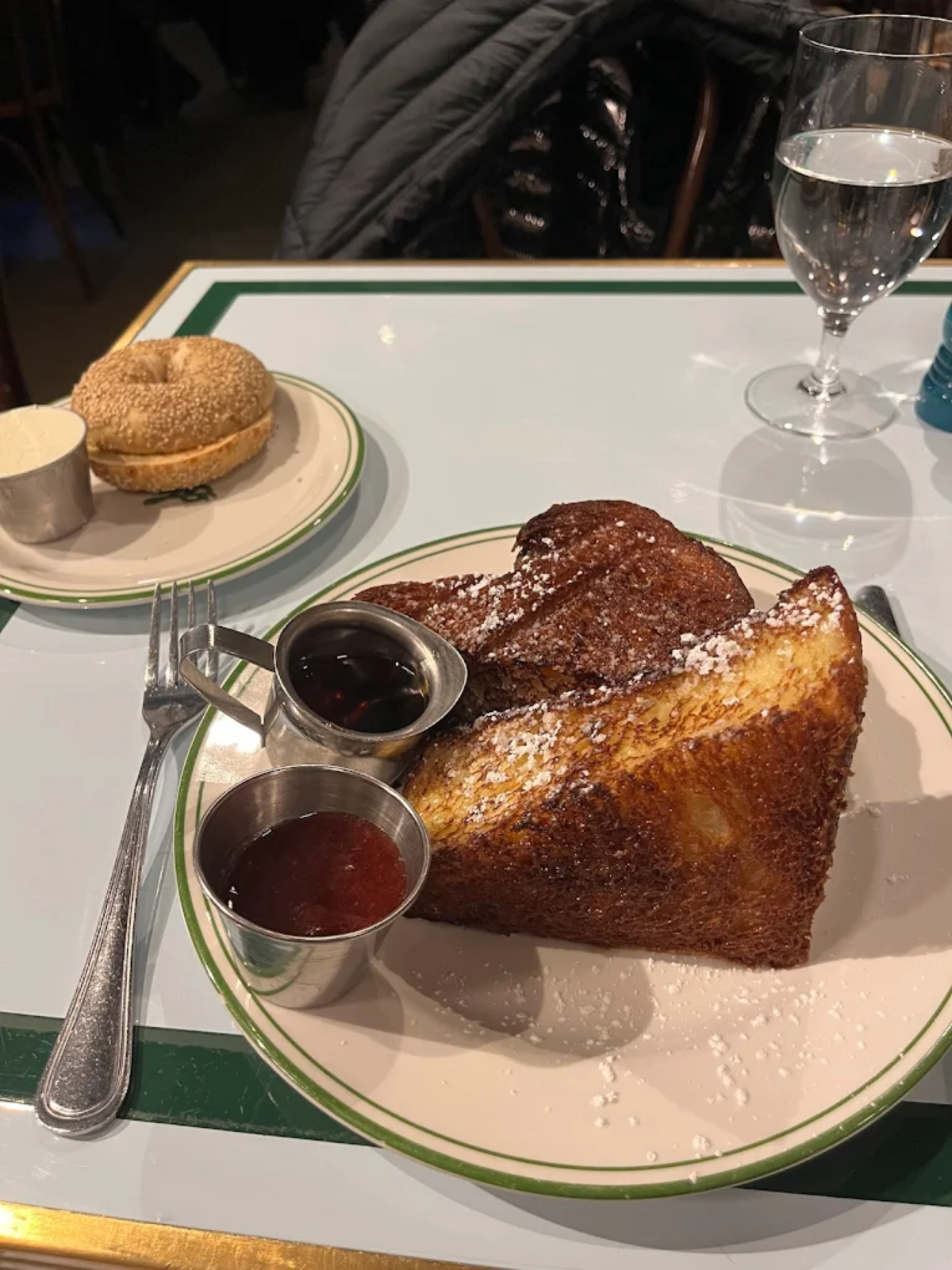 A plate with thick slices of French toast dusted with powdered sugar, served with two small cups of syrup and jam. Next to it is a plate with a plain bagel and a small dish of cream cheese, plus a glass of water.
