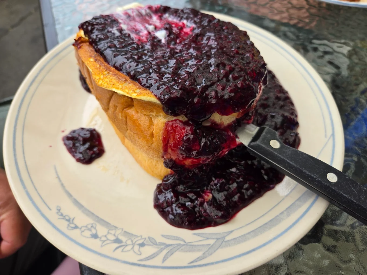 A thick slice of bread topped with a generous serving of dark berry jam, overflowing onto the plate, with a knife resting on the side. The plate has a light blue floral design around the rim.