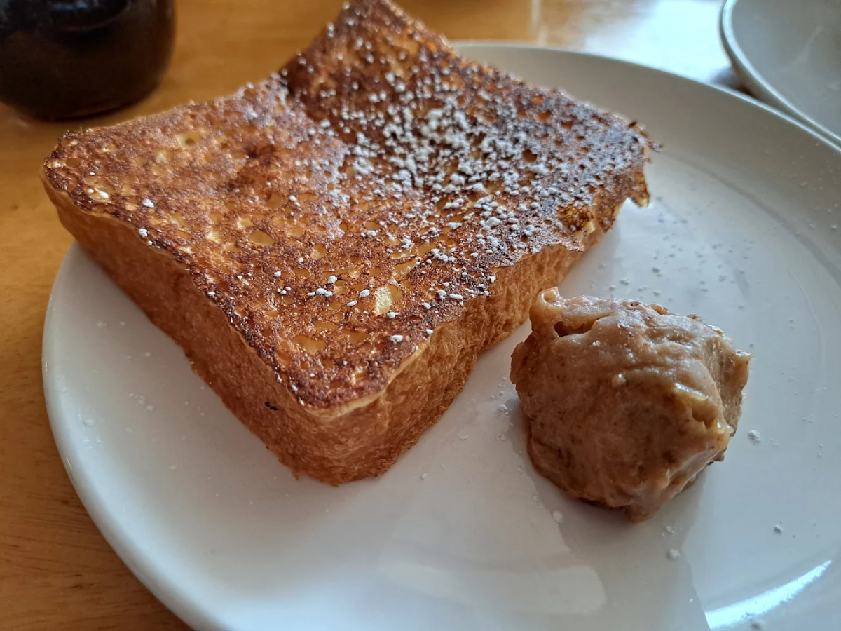 A thick slice of golden-brown French toast dusted with powdered sugar sits on a white plate next to a dollop of cinnamon-spiced butter.