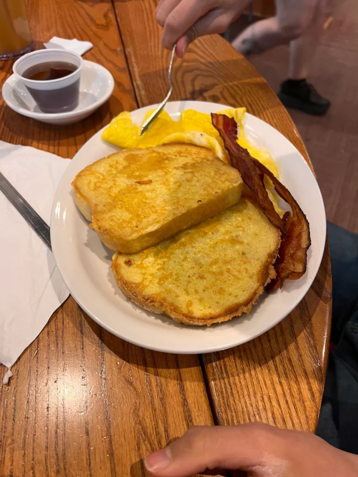 A plate with two slices of French toast, two strips of bacon, and a folded omelet. A small cup of syrup is on the side, and a hand is holding a fork near the eggs.