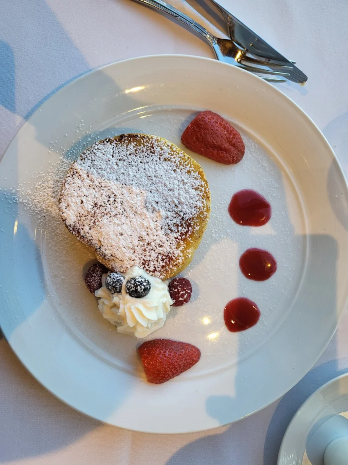 A dessert plated to resemble a bear's face, with a round pastry dusted with powdered sugar, whipped cream, berries for features, and strawberry halves with sauce dots for decorations, on a white plate.