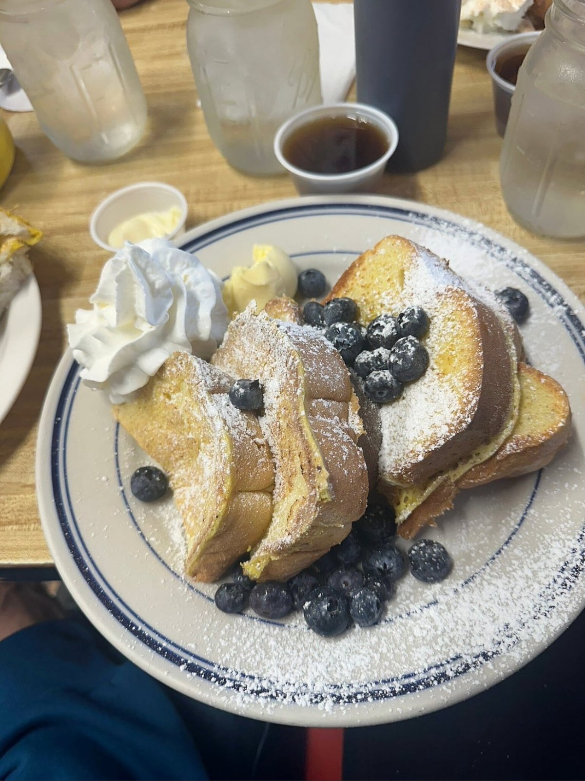 A plate of French toast topped with powdered sugar, fresh blueberries, whipped cream, and butter. Two mason jars with drinks, syrup, and small condiment cups are in the background on a wooden table.