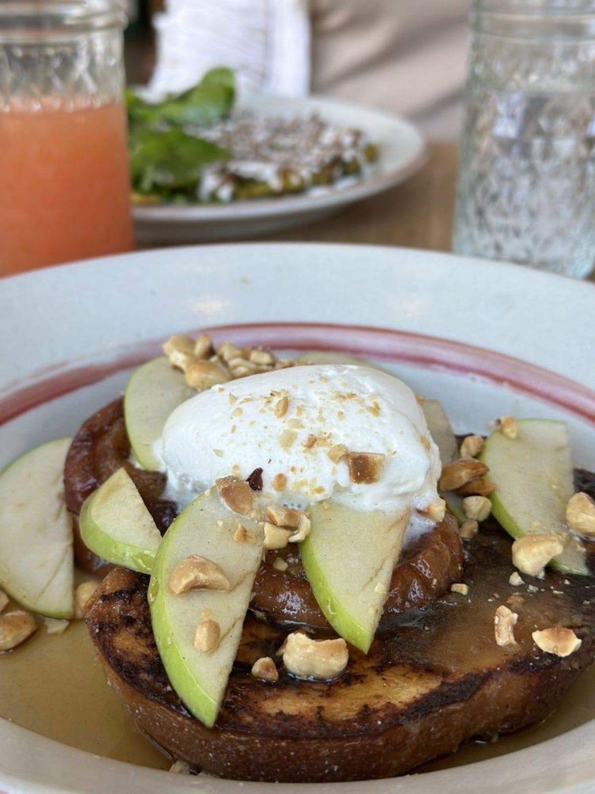 A close-up of French toast topped with green apple slices, chopped nuts, syrup, and a dollop of whipped cream. In the background are two drinks and a plate of food, slightly out of focus.
