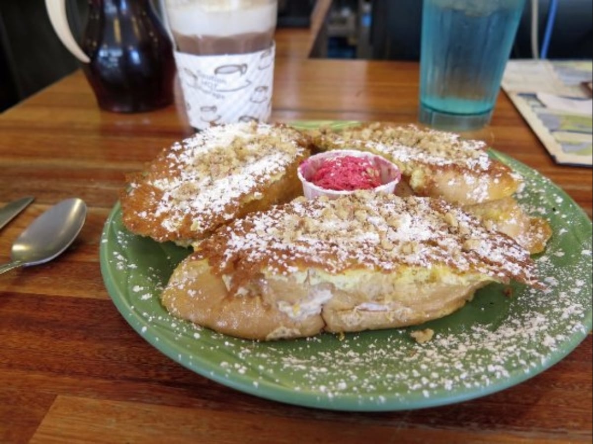 Three slices of French toast topped with powdered sugar and crushed nuts, served on a green plate with a small cup of pink spread, a spoon and fork nearby, and drinks in the background.
