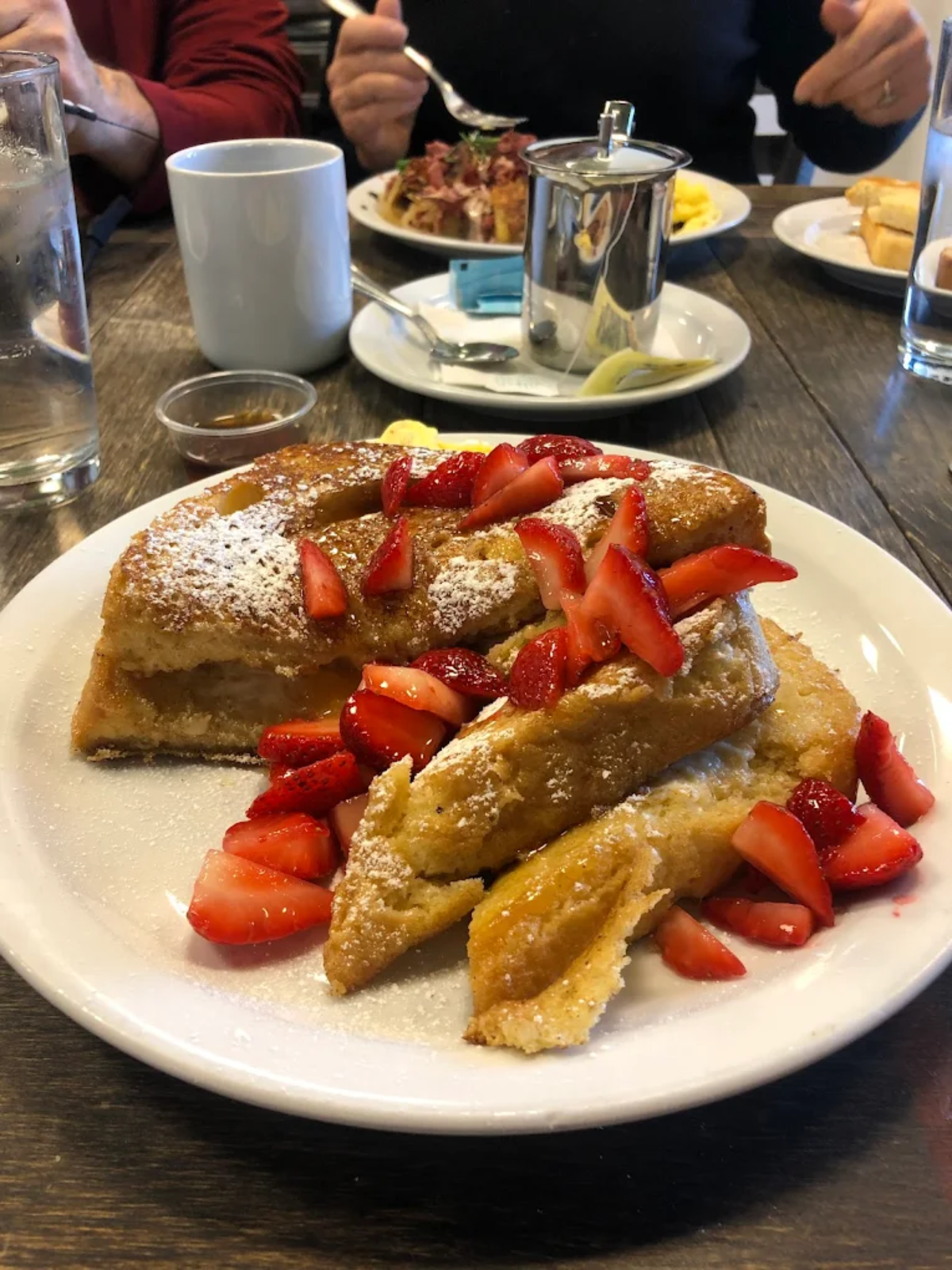 A plate of French toast topped with sliced strawberries and powdered sugar sits on a wooden table, with coffee, water, and other breakfast dishes visible in the background.