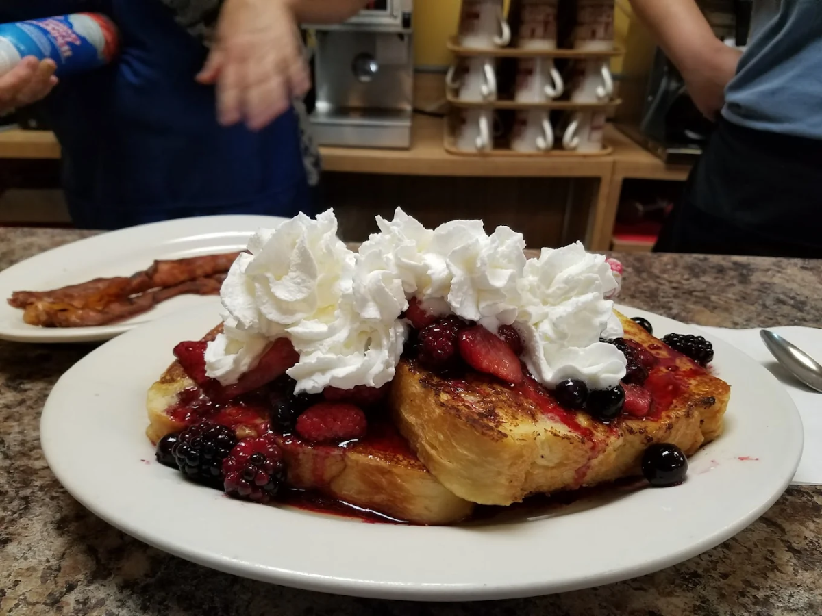 A plate of French toast topped with mixed berries and whipped cream sits on a counter, with a plate of bacon in the background and two people working behind the counter.