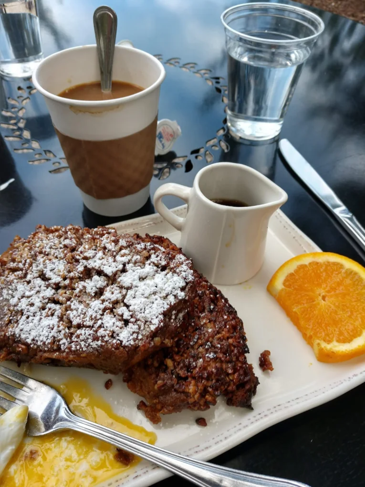 A plate with two slices of French toast topped with powdered sugar, orange slices, a small pitcher of syrup, and a cup of coffee sit on a glossy black table with a glass of water in the background.