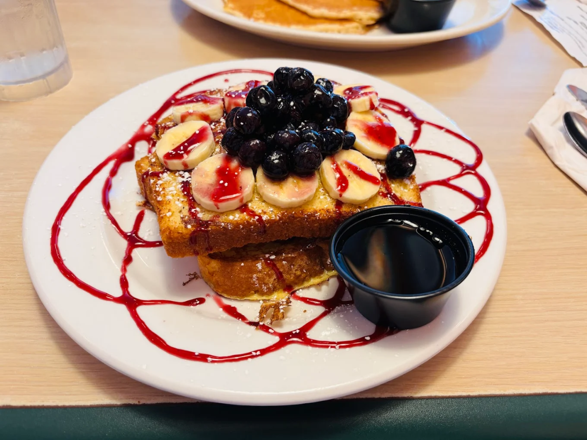 French toast topped with sliced bananas, blueberries, and berry sauce, dusted with powdered sugar, served with a side of syrup on a white plate. Another plate and glass of water are in the background.