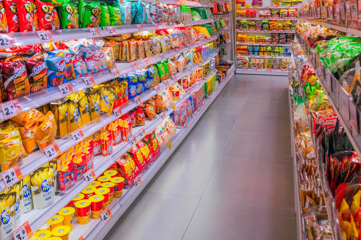 A brightly lit supermarket aisle with shelves stocked full of colorful snacks and chips on both sides, showcasing grocery store packaging tricks with eye-catching designs and price tags visible along the clear tile floor down the center.