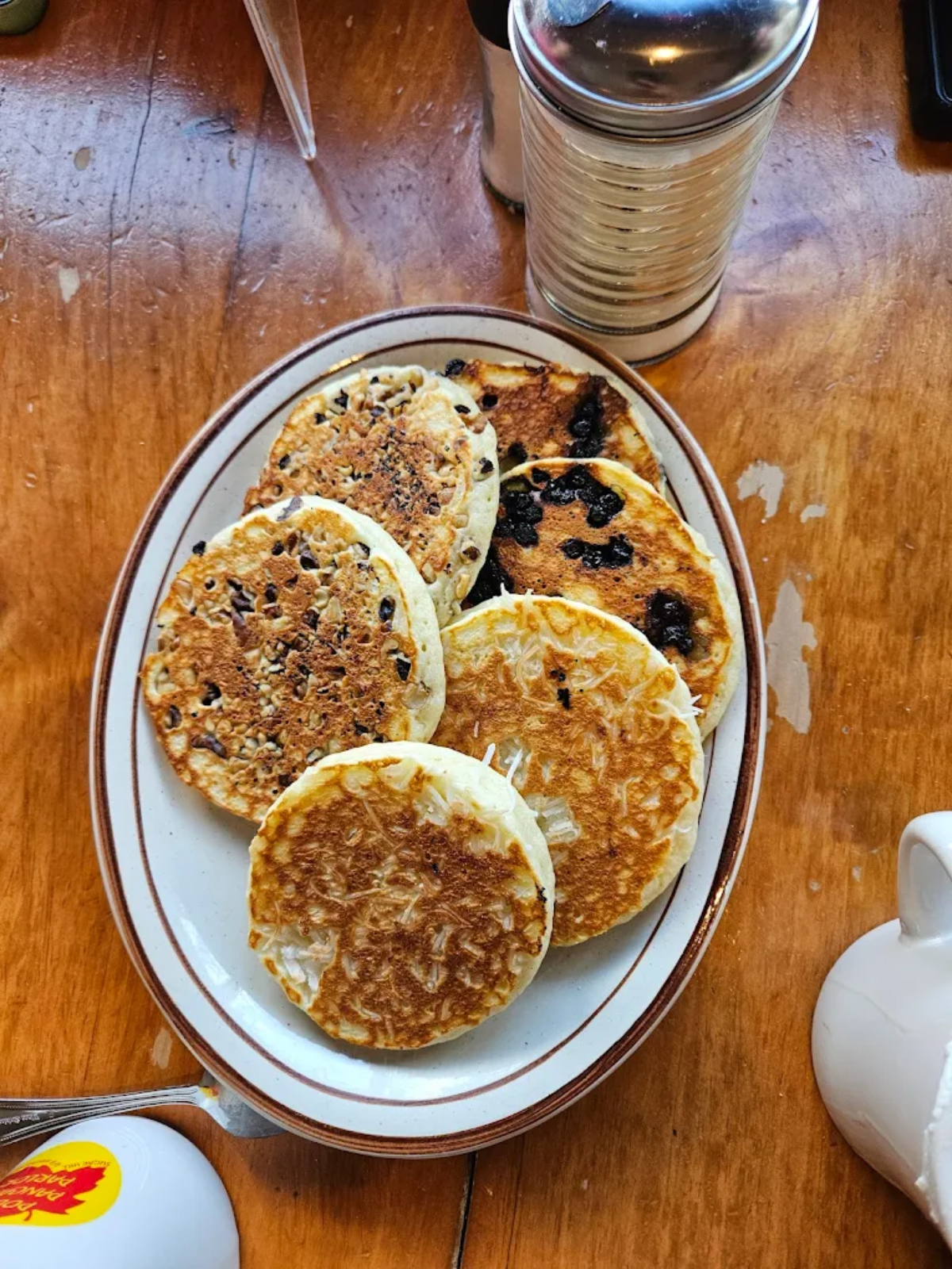 An oval plate holds six round pancakes, some with chocolate chips and some plain, on a wooden table next to a metal shaker and part of a syrup bottle.