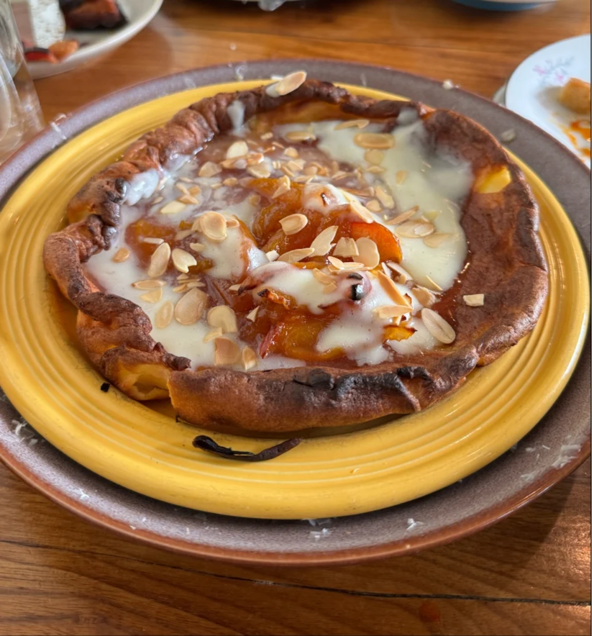 A golden-brown Dutch baby pancake topped with peach compote, melted cheese or cream, and sliced almonds sits on a yellow plate atop a larger brown plate on a wooden table.