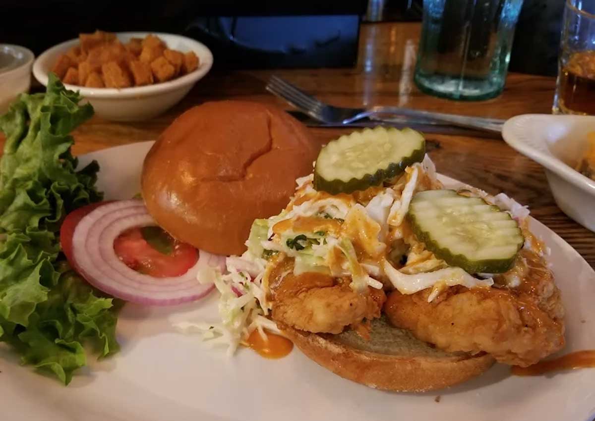 A fried chicken sandwich topped with coleslaw, pickles, and sauce on a bun, served with lettuce, tomato, red onion slices, and a side of croutons on a white plate.