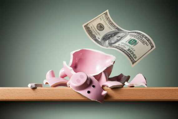 A broken pink piggy bank lies shattered on a wooden surface, with several pieces scattered around and a hundred-dollar bill floating above it against a muted green background.