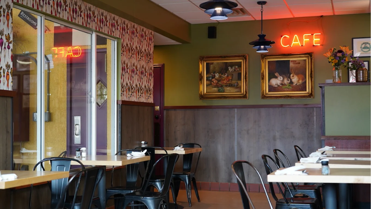 A cozy cafe interior with wooden tables, black chairs, neon "CAFE" sign on the wall, two framed paintings, floral wallpaper, and a vase of flowers on a counter. Place settings are ready for customers.