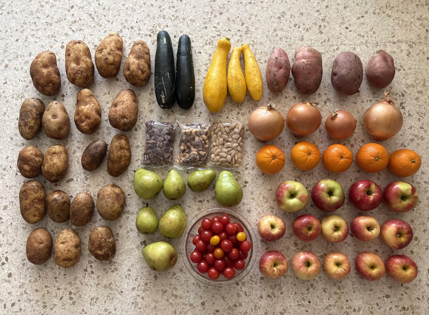 A variety of fruits, vegetables, and nuts arranged on a countertop: potatoes, zucchini, yellow squash, sweet potatoes, onions, oranges, pears, apples, cherry tomatoes in a bowl, and small bags of nuts and beans.