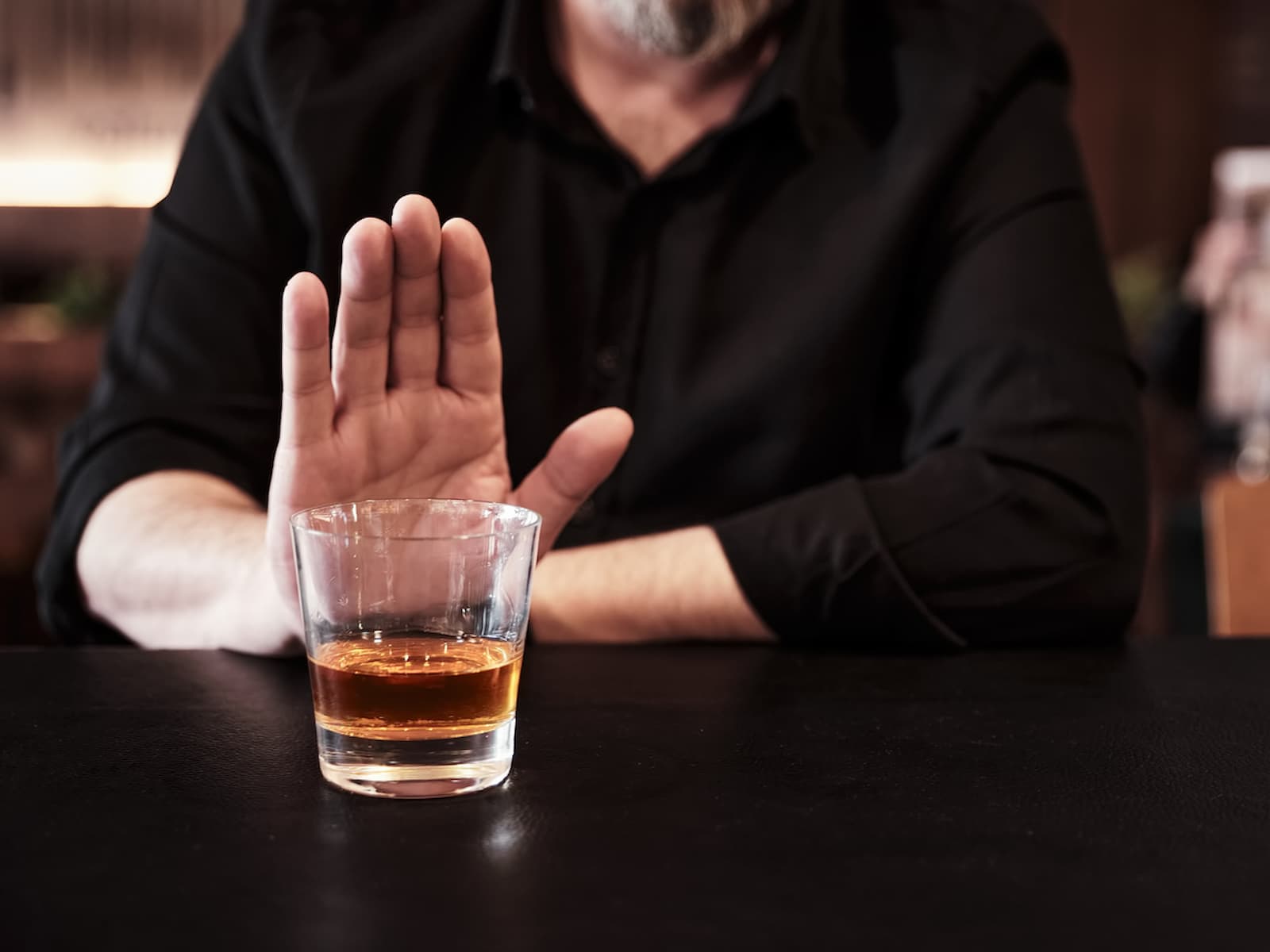 A person with a beard, wearing a black shirt, sits at a table and holds up their hand in refusal toward a glass of whiskey in front of them, demonstrating strong money habits and self-discipline.