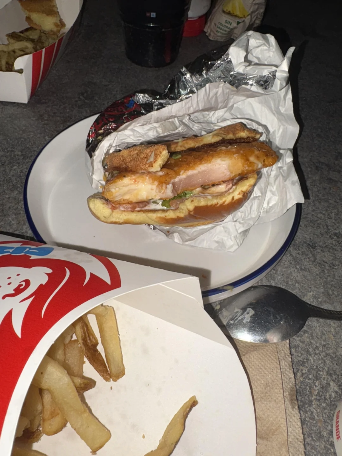 A partially unwrapped chicken sandwich sits on a plate, with a box of French fries and a metal spoon nearby on a napkin. The meal appears to be from a fast food restaurant.