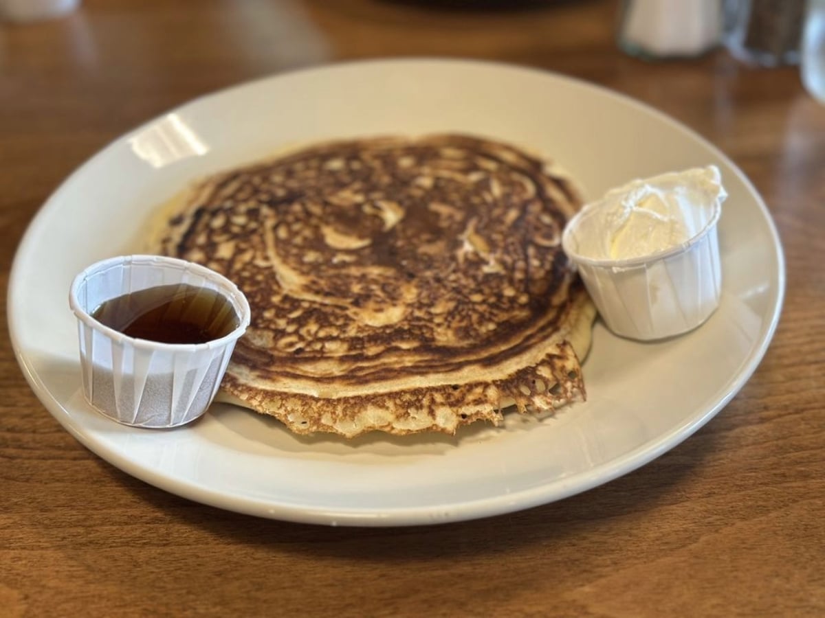 A golden-brown pancake on a white plate, served with a small cup of syrup and a small cup of whipped butter on the side, sitting on a wooden table.