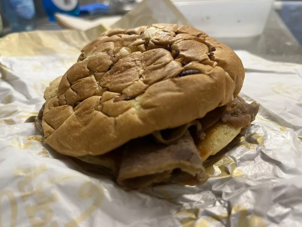 A close-up of a sandwich with a squished, wrinkled bun and slices of meat inside, resting on a sheet of branded sandwich paper. The background includes a blurred water bottle and food container.