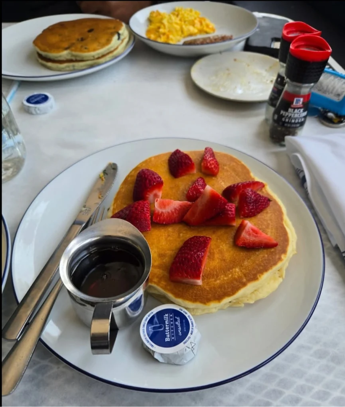 A plate with pancakes topped with sliced strawberries, a cup of syrup, and a packet of butter. Knife and fork rest on the plate. In the background are another plate with pancakes and a plate with scrambled eggs and sausage.