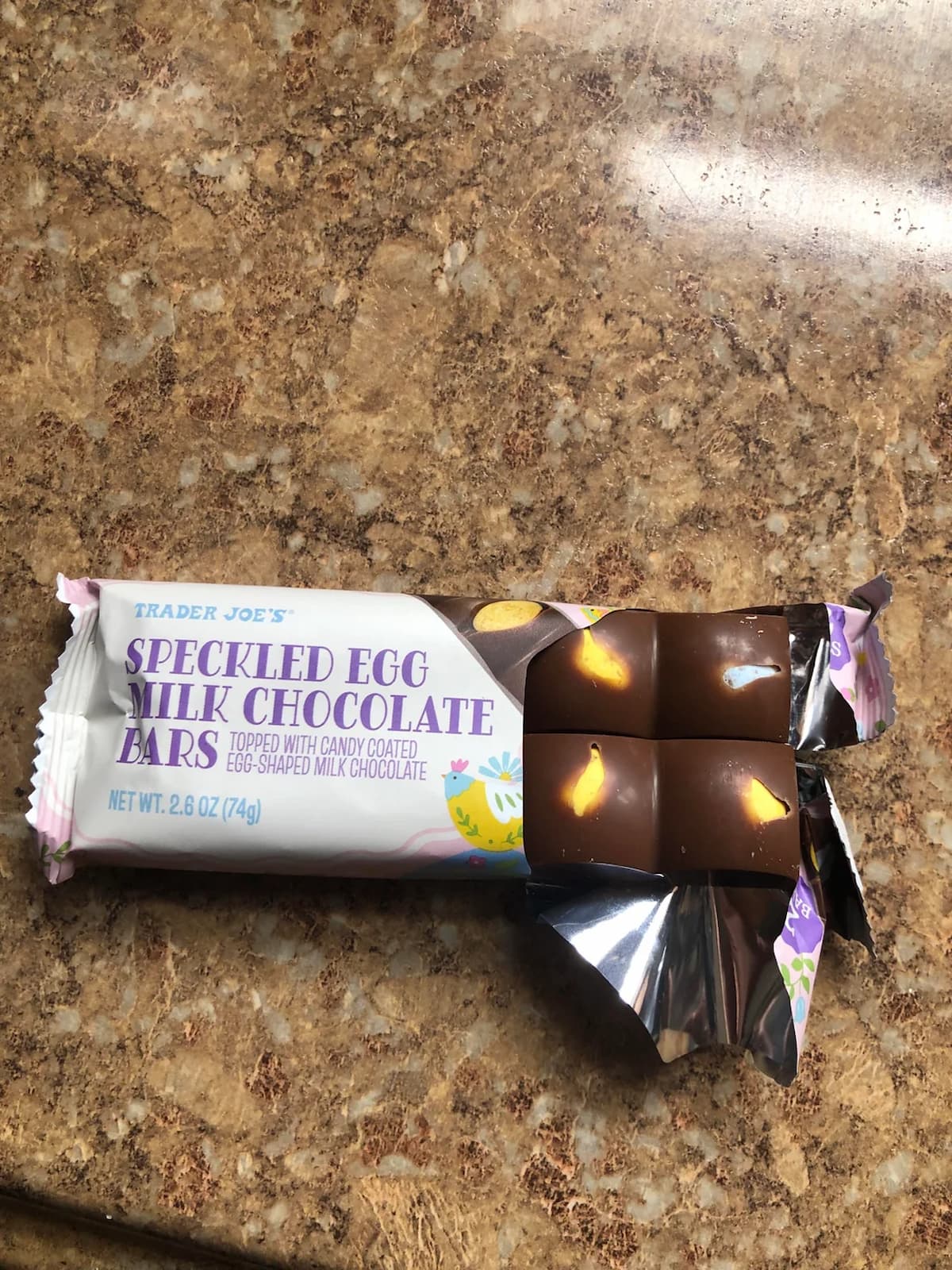 A partially unwrapped Trader Joe’s Speckled Egg Milk Chocolate Bar sits on a brown countertop, showing four chocolate squares with yellow and white speckled candy pieces inside.