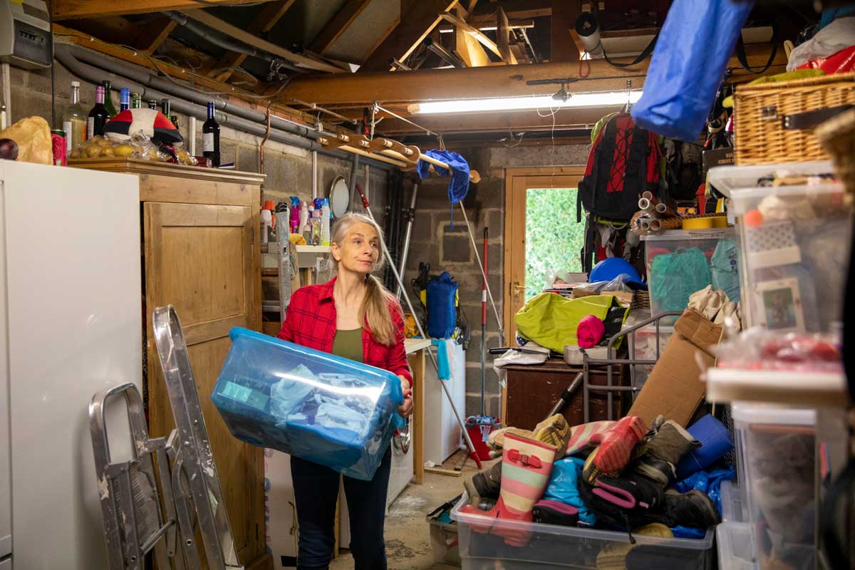 A woman in a red plaid shirt holds a large blue storage bin in a cluttered garage filled with boxes, tools, and things to toss for spring cleaning. She looks up, appearing to organize or rearrange the space.