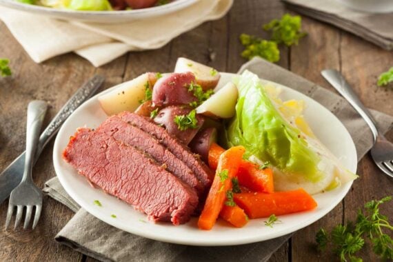 A classic St. Patrick's Day dinner: corned beef with sliced carrots, potatoes, and cabbage on a white plate, garnished with parsley, set on a rustic wooden table with utensils nearby.