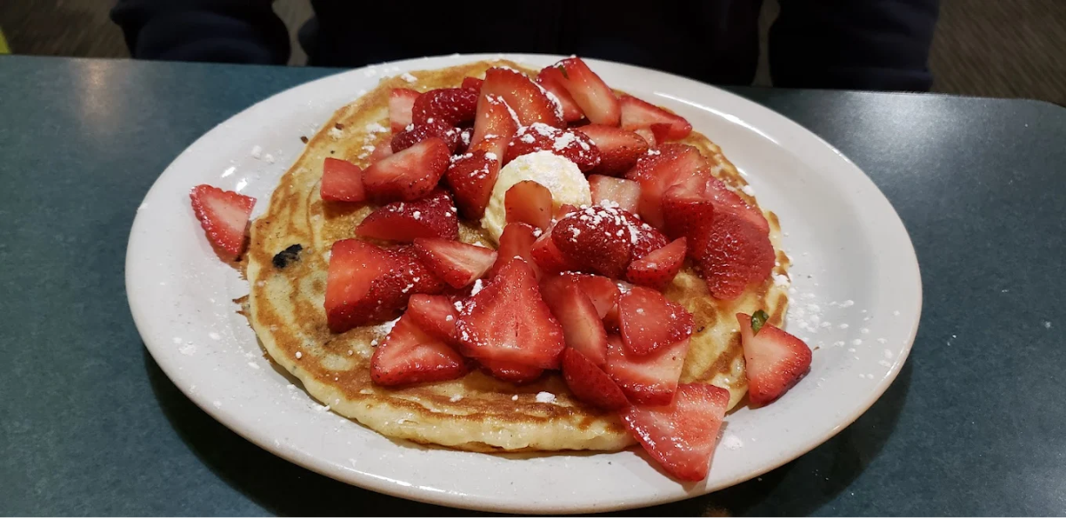 A plate with a large pancake topped with sliced strawberries, a dollop of butter, and a dusting of powdered sugar sits on a green table.