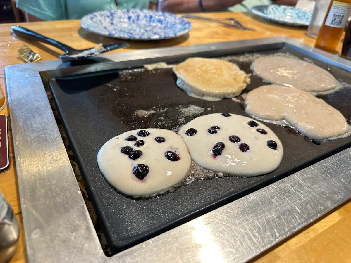Pancakes are cooking on a griddle, with two featuring blueberries and three plain. Plates and utensils are visible in the background, suggesting a breakfast setting.