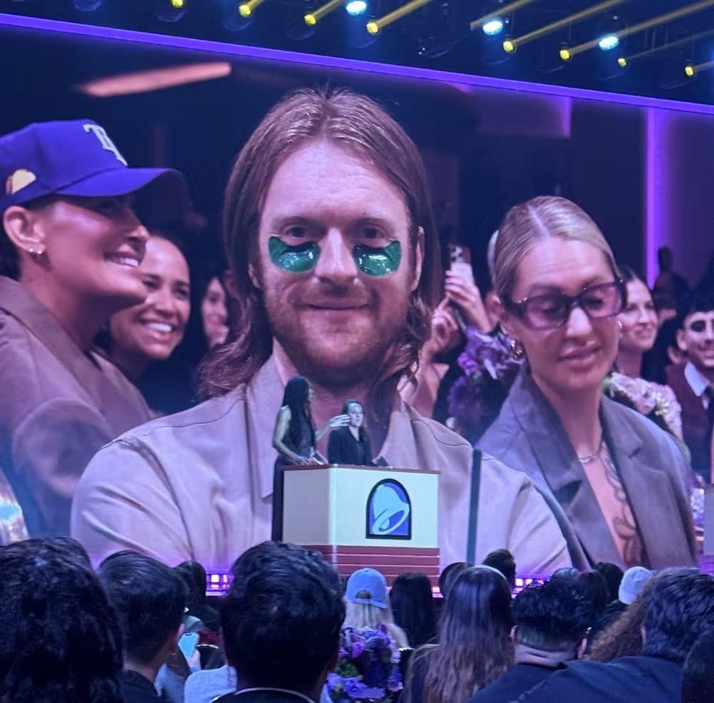 A man with long hair and green under-eye patches is shown on a large screen at an indoor event, standing among a crowd. People around him are smiling and the atmosphere appears festive.