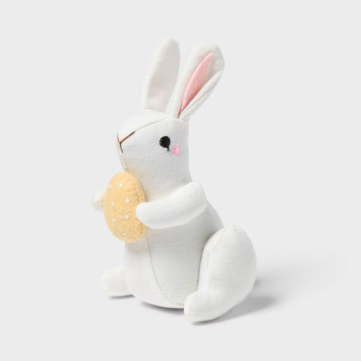 A white plush bunny toy with pink-lined ears and a small pink cheek, sitting upright and holding a tan, round object with white dots, against a plain light background.