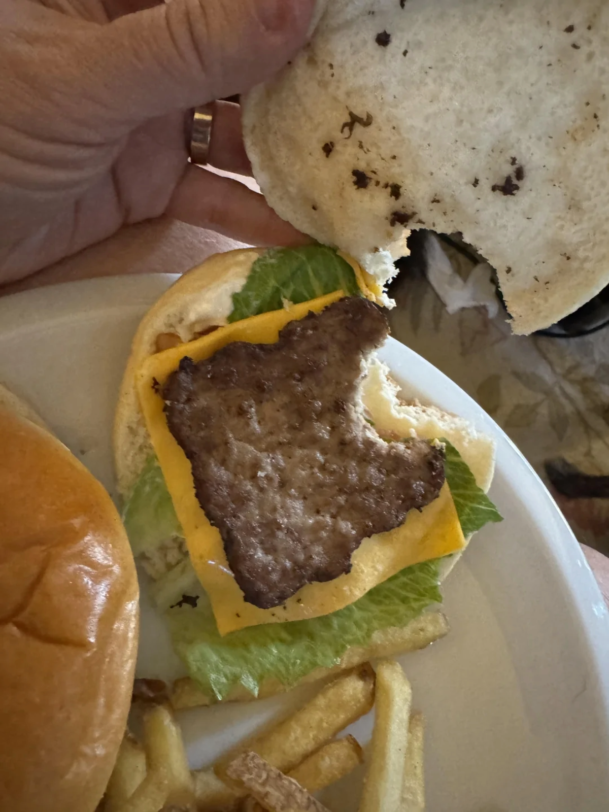 A hand lifts the top bun of a burger, revealing a small, uneven patty on a slice of cheese and lettuce. The plate also holds fries and another sandwich or bun.