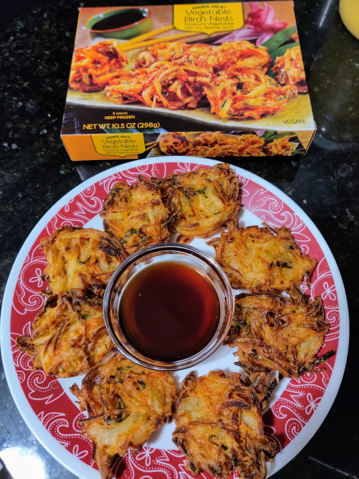 A plate of crispy vegetable fritters arranged in a circle around a small bowl of dipping sauce, with the packaging for "Vegetable Bird's Nests" in the background on a dark countertop.