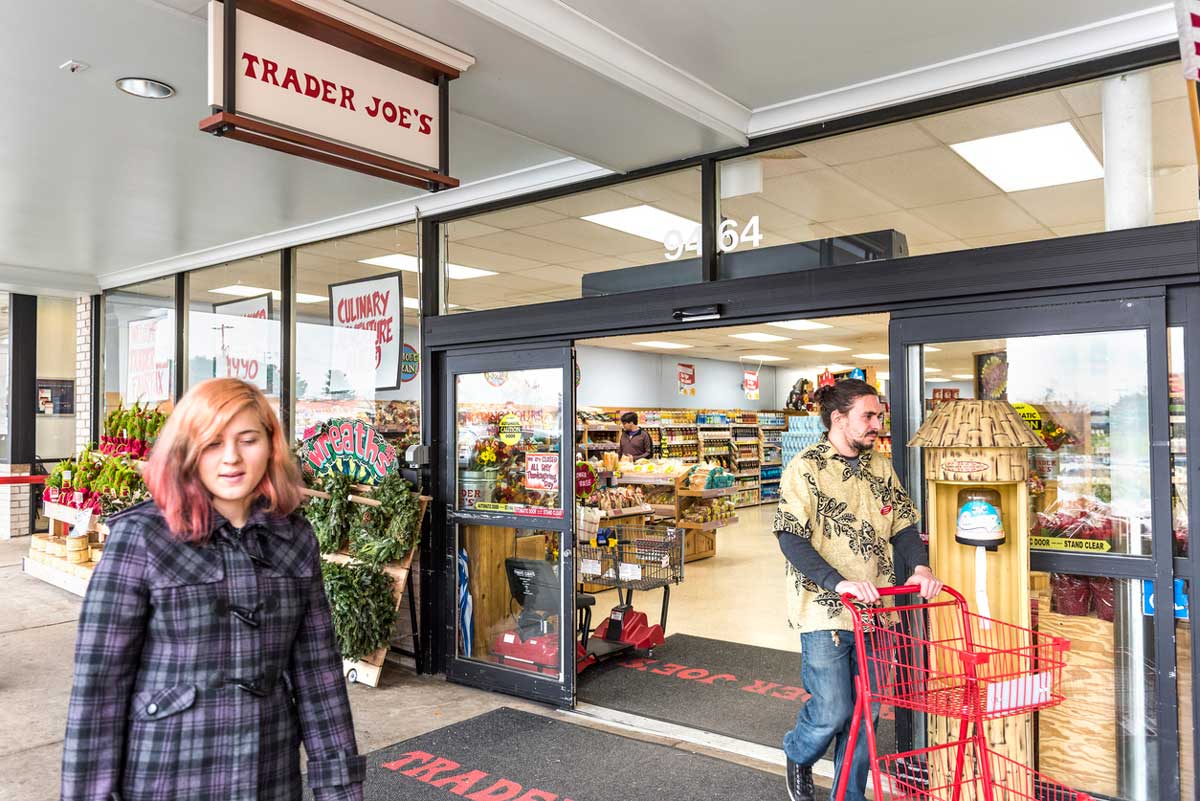 A woman walks past the entrance of a Trader Joe’s store as a man pushes a red shopping cart out the automatic doors. Displays of plants and grocery items are visible outside and inside the store.