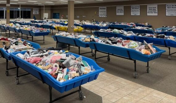 Large room filled with rows of blue plastic bins on wheels, each overflowing with assorted household items, toys, and goods. Signs hang on the walls, and the floor is a mix of carpet and tile.