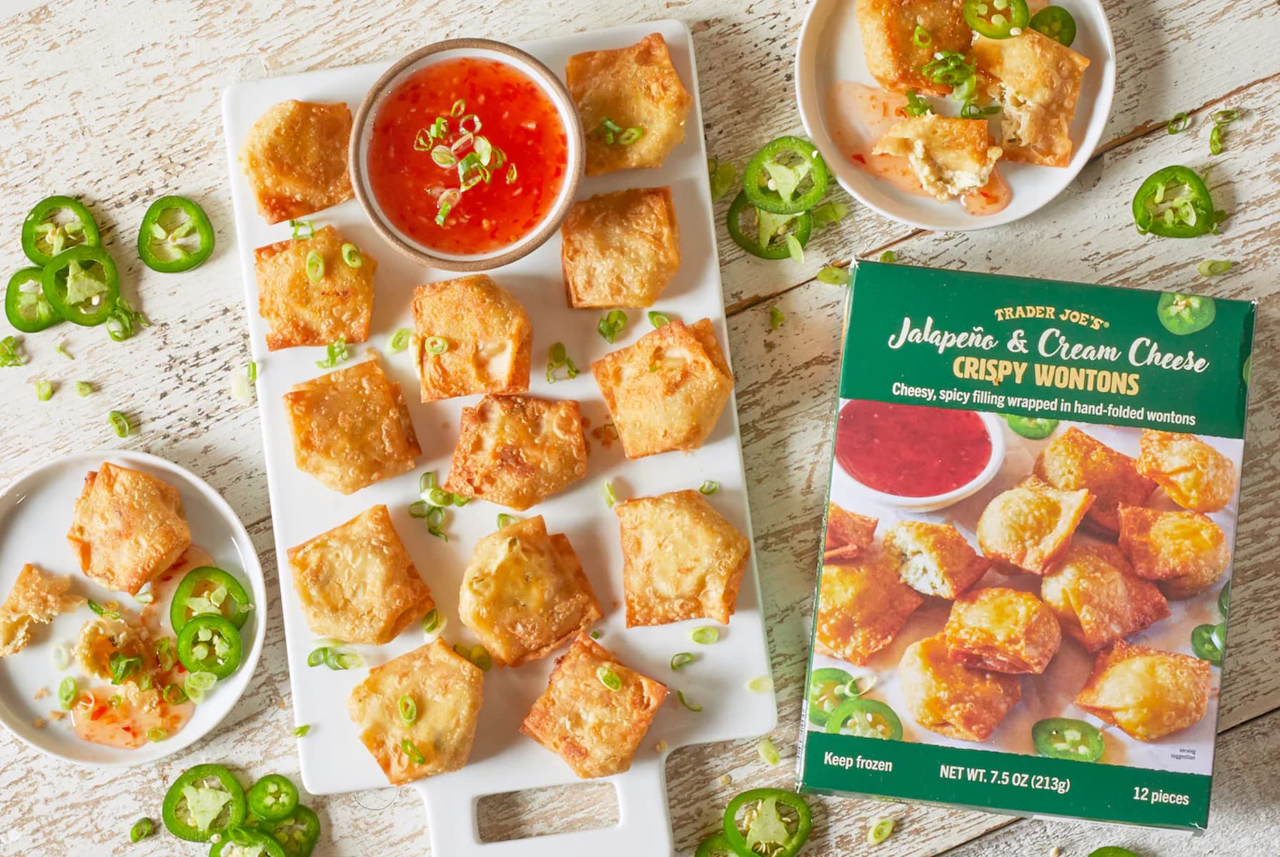 A platter of golden, crispy wontons garnished with green onions, served with a bowl of red dipping sauce. Sliced jalapeños and an open Trader Joe’s Jalapeño & Cream Cheese Crispy Wontons box are nearby.