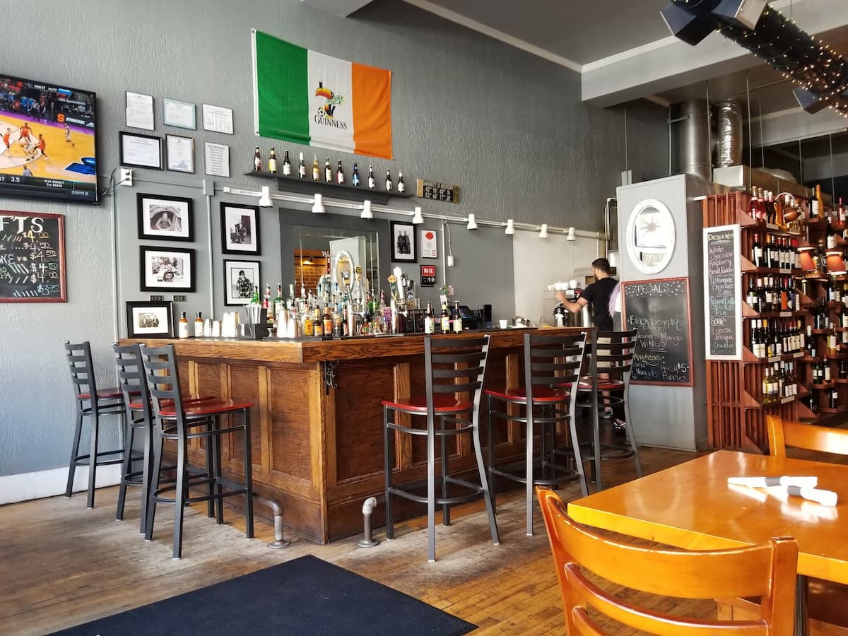 A cozy bar with a wooden counter surrounded by high chairs, liquor bottles on display, framed photos on gray walls, an Irish flag overhead, and shelves of wine bottles on the right. A TV shows a sports game.