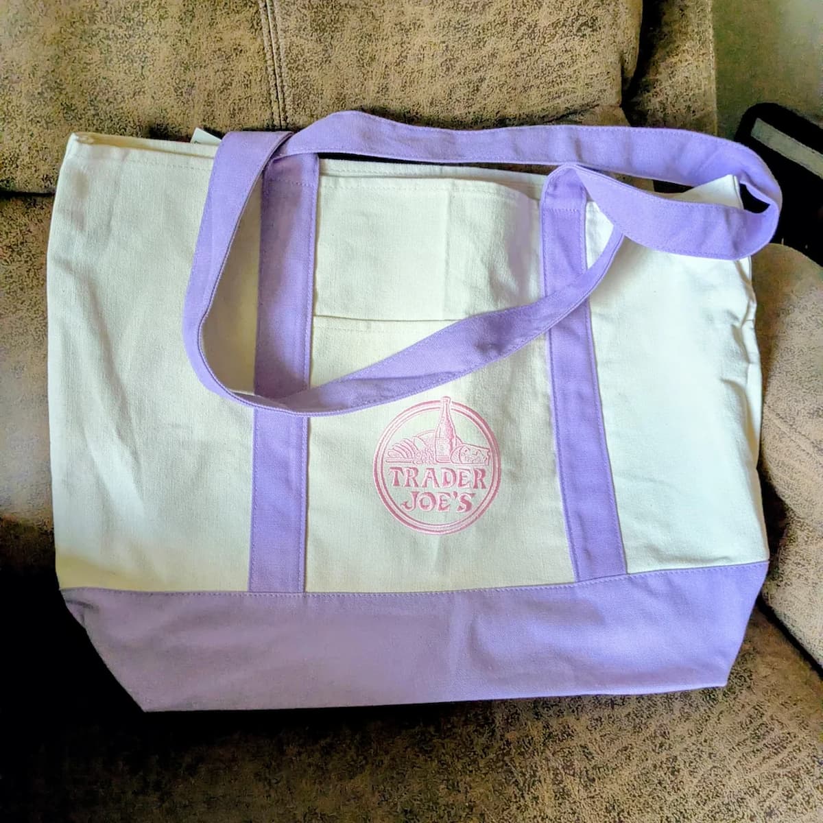 A cream and lavender canvas tote bag with purple straps sits on a tan couch. The bag features the Trader Joe's logo in purple on the front center.
