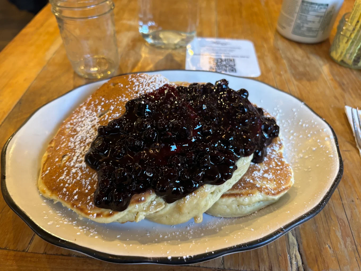 A plate of two fluffy pancakes topped with a generous serving of blueberry compote and a dusting of powdered sugar, on a wooden table with glass jars and a water bottle in the background.